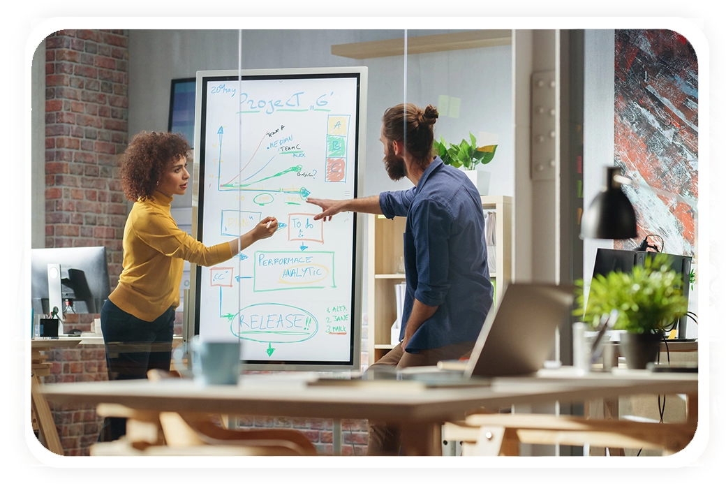 A woman and a man discuss a project at a whiteboard filled with diagrams and notes in a modern office setting.